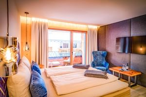 Chambre double moderne avec literie blanche, fauteuil bleu, TV murale et portes-fenêtres donnant sur terrasse avec vue montagne.