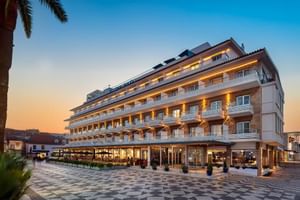 Modernes weißes Hotel Baia Gebäude mit mehreren Balkonen und warmer Beleuchtung bei Sonnenuntergang. Gemusterter Steinplatz im Vordergrund mit Palmen.