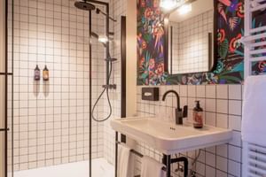 Hotel Labé Tours Badezimmer mit weißen Fliesen, Glasdusche, schwarzen Armaturen und bunter Blumentapete um den Spiegel über dem Waschbecken.