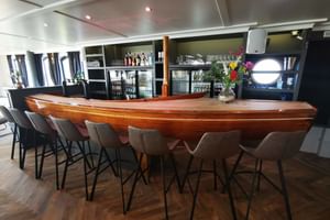 Moderne Restaurant-Bar auf der MS Magnifique IV mit geschwungener Holztheke, grauen Barhockern und Glaskühlschränken mit Getränken.