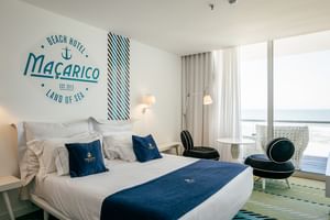 Modernes Doppelzimmer im Macarico Beach Hotel mit weißer Bettwäsche, blauen Akzenten, nautischer Wanddekoration und großen Fenstern mit Meerblick.