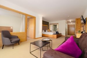 Moderne Hotelsuite im Mein Almhof mit braunem Sofa, pinkem Kissen, grauem Sessel, schwarzem Couchtisch und hölzernem Schlafbereich.