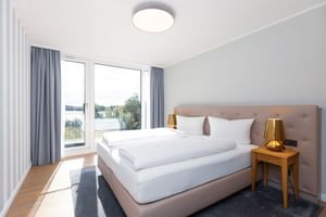 Helles Hotelzimmer mit großem Bett, grauen Vorhängen, Holznachttischen mit goldenen Lampen und Balkontür mit Blick auf die Müritz.