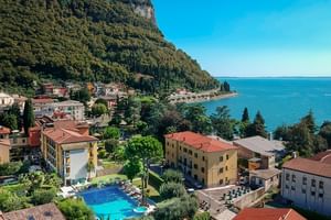 Panoramaansicht vom Hotel Regina Adelaide mit bunten Gebäuden, Schwimmbad, blauen Gewässern des Gardasees und bewaldeten Bergen.