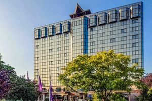 Modernes Mercure Hotel in Koblenz mit Glasfassade und blauen Akzenten, umgeben von grünen Bäumen und Grünanlagen unter blauem Himmel.