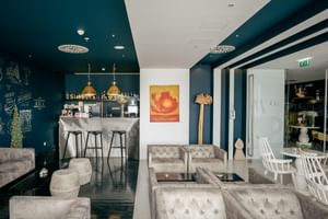 Moderne Hotelbar mit türkisfarbener Decke, weißen gesteppten Sitzgelegenheiten, Messingpendelleuchten und Bartheke mit Flaschen.