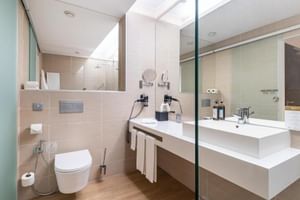 Modernes Badezimmer im Hotel Jupiter Lisboa mit weißem Waschtisch, Wand-WC, Glasdusche und beigen Fliesen mit Holzboden.