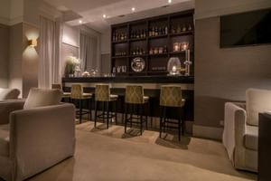 Moderne Hotelbar mit dunkler Holztheke, vier grünen Barhockern mit Rautenmuster, Flaschenregalen und beigem Sitzbereich mit Sesseln.