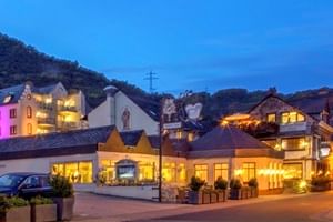 Abendansicht des Schloss Hotel Petry in Trais-Karden mit beleuchteten weißen Gebäuden, Kapelle und bewaldeten Hügeln unter blauem Himmel.