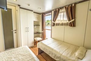 Helle Zweibettkabine auf der MS Olmissum mit zwei Betten, cremefarbenen Wänden, Holzboden, Schrank, Waschtisch und Fenster mit braunen Vorhängen.