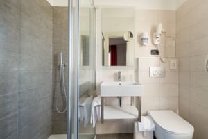 Helles Bad mit beigen Fliesen, Glasdusche, Wandwaschbecken, Spiegel und weißer Toilette. Pflegeprodukte und Handtücher vorhanden.