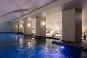 Moderner Indoor-Spa-Pool mit blauer Beleuchtung im Hotel Fleur de Loire Blois mit weißen Säulen, Liegestühlen und Deckenbeleuchtung.