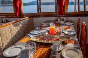 Frühstückstisch auf der MS Macek mit Tellern, Tassen, Saft und Aufschnitt. Große Fenster mit roten Vorhängen zeigen Seeblick mit bewaldeten Ufern.