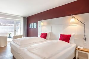 Modernes Superior Hotelzimmer mit zwei Betten, weißem Kopfteil, roten Kissen, bordeauxroter Akzentwand und Seeblick durch große Fenster.