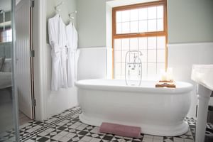 Weiße freistehende Badewanne in hellem Bad mit geometrischen Bodenfliesen, weißen Bademänteln an Haken und Schiebefenster. Unterkunft Nordirland.