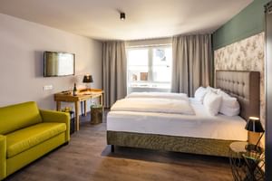 Modernes Doppelzimmer Superior mit grüner Akzentwand, grau gepolstertem Bett, lindgrünem Sofa, Holzschreibtisch, Flachbildfernseher und großem Fenster.