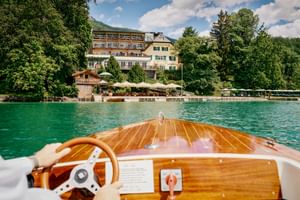 Blick vom Holzboot auf das Landhaus zu Appesbach am türkisfarbenen See. Hotelgebäude mit Terrasse und Sonnenschirmen am Ufer sichtbar.