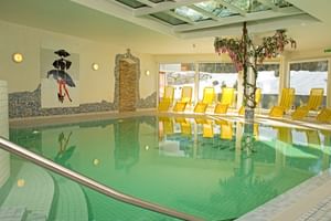 Hallenschwimmbad mit türkisfarbenem Wasser, gelben Liegestühlen entlang der Fenster, Wanddekoration und Glasdeckenpaneelen im Hotel.