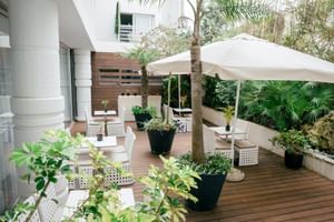 Holzterrasse im Macarico Beach Hotel mit weißen Korbmöbeln, großem Sonnenschirm, Topfpflanzen und tropischer Vegetation.