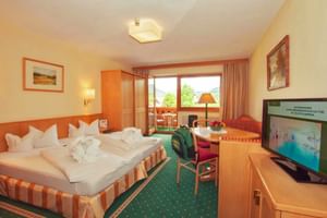 Gemütliches Hotelzimmer mit Doppelbett, orange gestreifter Bettwäsche, grünem Teppich, Holzmöbeln, TV, Balkon mit Bergblick und traditioneller Alpenausstattung.