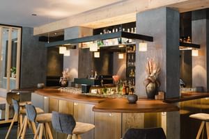Moderner Barbereich im Mein Almhof mit Betonsäulen, Holztheke, Hängeleuchten und Barhockern. Flaschen und Dekovasen sind sichtbar.