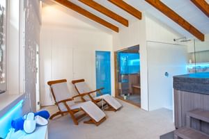 Espace bien-être moderne avec poutres en bois, deux chaises longues, sauna avec porte vitrée, douche et jacuzzi. Murs blancs et éclairage ambiant bleu.