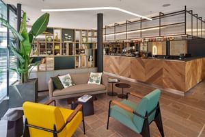 Zeitgenössische Hotelbar mit Holztheke, buntem Sitzbereich mit gelben und türkisfarbenen Stühlen, grauem Sofa und großer Pflanze.
