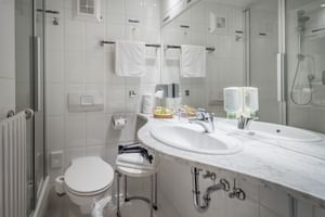 Modernes weiß gefliestes Badezimmer im Salzburger Hof Bad Gastein mit Waschbecken, Spiegel, Dusche, WC und Handtüchern. Amenities auf der Ablage.