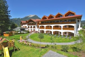 Großes Alpenhotel mit weißer Fassade und Holzbalkonen in grüner Anlage mit Spielplatz, umgeben von Bergen unter blauem Himmel.