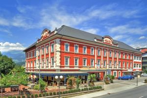 Hotel Ertl Spittal, ein rotes dreistöckiges Gebäude mit weißen Verzierungen und grauem Dach. Erdgeschoss-Terrasse mit Außenbestuhlung unter blauem Himmel.