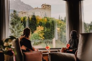 Zwei Frauen sitzen in Hotelsesseln mit Getränken und blicken durch große Fenster auf eine mittelalterliche Burg umgeben von Herbstwald und Bergen.