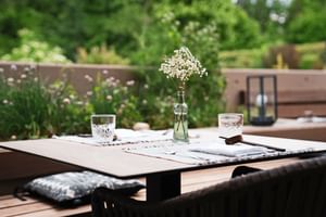 Holztisch auf der Terrasse im Bergrose Hideaway mit zwei Gläsern, weißen Blumen in Glasvase, Servietten und Kissen, umgeben von üppigem Grün.