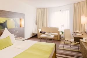 Modernes Doppelzimmer im Mercure Koblenz mit weißem Bett, grünen Akzenten, beigem Sofa, Schreibtisch und großen Fenstern mit Vorhängen.