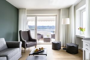 Modernes Wohnzimmer mit grauen Sesseln, Sofa und Couchtisch. Große Fenster mit weißen Vorhängen öffnen zum Balkon mit See- und Bergblick.