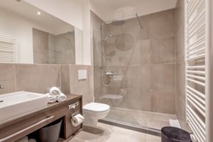 Modernes Badezimmer im Hotel Ertl in Spittal mit beigen Fliesen, Glasdusche, weißem Waschbecken, WC, Handtuchheizkörper und Handtüchern.