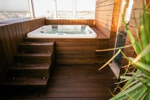 Weißer Whirlpool in Holzdeck mit Stufen im Macarico Beach Hotel. Große Fenster zeigen Küstenblick mit grüner Pflanze im Vordergrund.