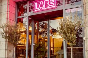 Moderner Glaseingang des Hotel Labé mit pinker Neonbeschilderung über der Tür. Zwei Olivenbäume in weißen Töpfen flankieren den Eingang.