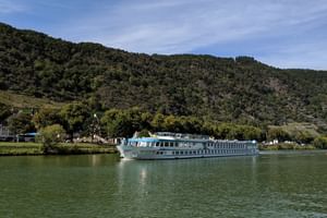 Weißes Flusskreuzfahrtschiff Switzerland auf ruhigem grünen Wasser mit bewaldeten Hügeln und kleinem Dorf im Hintergrund unter blauem Himmel.