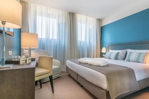 Modernes Doppelzimmer im Universal Boutique Hotel mit blauer Akzentwand, weißem Bett, gestreiften Kissen, Schreibtisch mit Lampe und Sessel am Fenster.