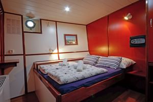 Doppelkabine auf der MS Macek mit Holzbett, gestreiften Kissen, Bullauge und warmer Holzvertäfelung. Ein Safe ist an der roten Wand montiert.