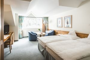 Chambre double Gampen lumineuse avec lits jumeaux, fauteuils bleus, mobilier en bois, TV et grande fenêtre avec rideaux turquoise.