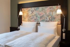 Modernes Hotelzimmer mit weißer Bettwäsche, buntem geometrischem Kopfteil, verstellbaren Leselampen und dunkler Akzentwand im Alte Post Konstanz.