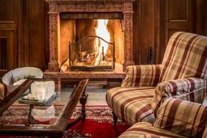 Eleganter Lobby-Kaminbereich im Regina Adelaide Garda mit verziertem Steinkamin, brennendem Feuer, karierten Sesseln und Perserteppich.