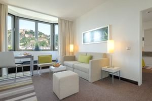 Moderne Hotellounge im Mercure Koblenz mit grauem Sofa, grünen Kissen, weißem Hocker, Stehlampen und großen Fenstern mit Stadtblick.