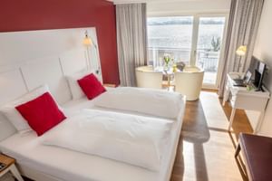 Modernes Hotel-Doppelzimmer mit weißem Bett, roten Akzentkissen, Sitzbereich am Fenster und Seeblick in der Residenz Seeterrasse Wangen.