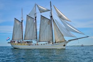 Dreimaster-Segelschiff Mare Frisium mit weißen Segeln auf ruhigem blauen Wasser unter klarem Himmel. Niederländische Flagge am Heck sichtbar.