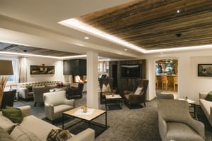 Salon d'hôtel moderne avec fauteuils gris, poutres de plafond en bois, cheminée et bar. Mobilier contemporain arrangé en groupes.