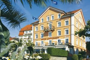 Gelbes Hotel Reutemann Gebäude in Lindau mit weißen Verzierungen, mehreren Stockwerken, Balkonen und Außenterrasse umgeben von Palmen und Blumen.