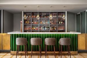 Moderne Hotel Baia Bar mit grün gefliester Theke, vier grauen Barhockern, umfangreicher Flaschenauslage in Regalen und Pendelleuchten.