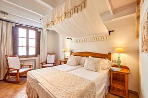 Chambre d'hôtel lumineuse à Pollença avec lit double, baldaquin blanc, mobilier en bois, sol en terre cuite et deux lampes de chevet.